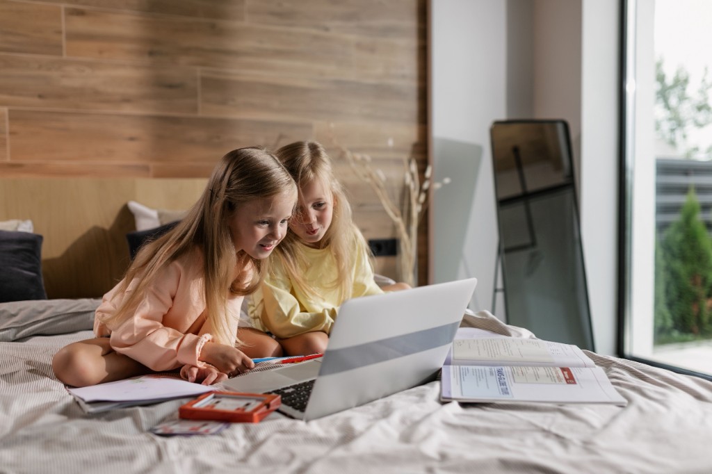 Two young girls learning together at home on a bed with a laptop.
